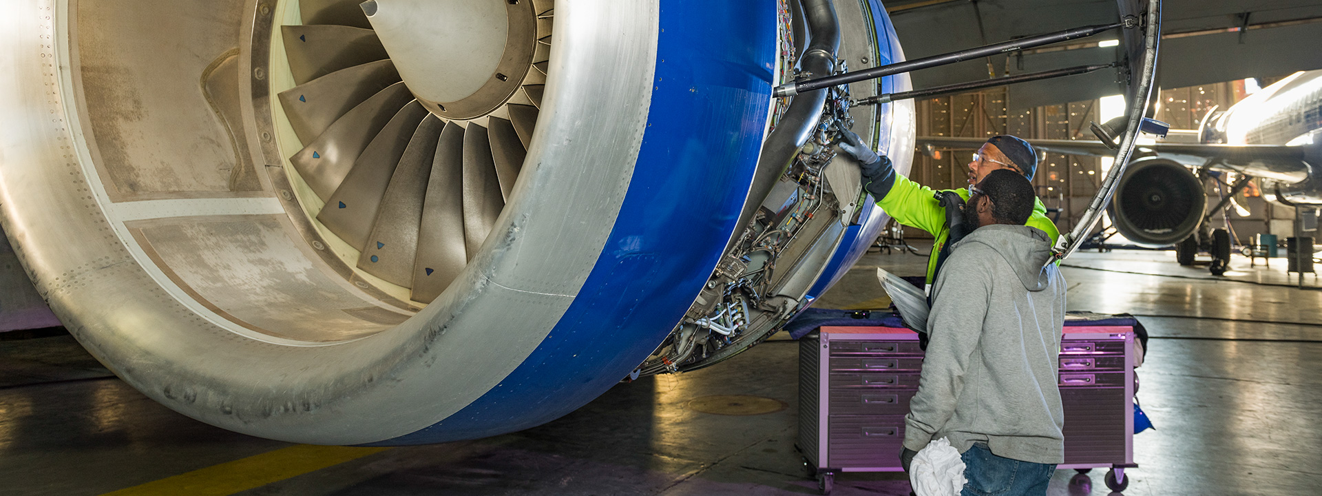 Aircraft maintenance technicians inspect a large jet engine inside an aircraft hangar.