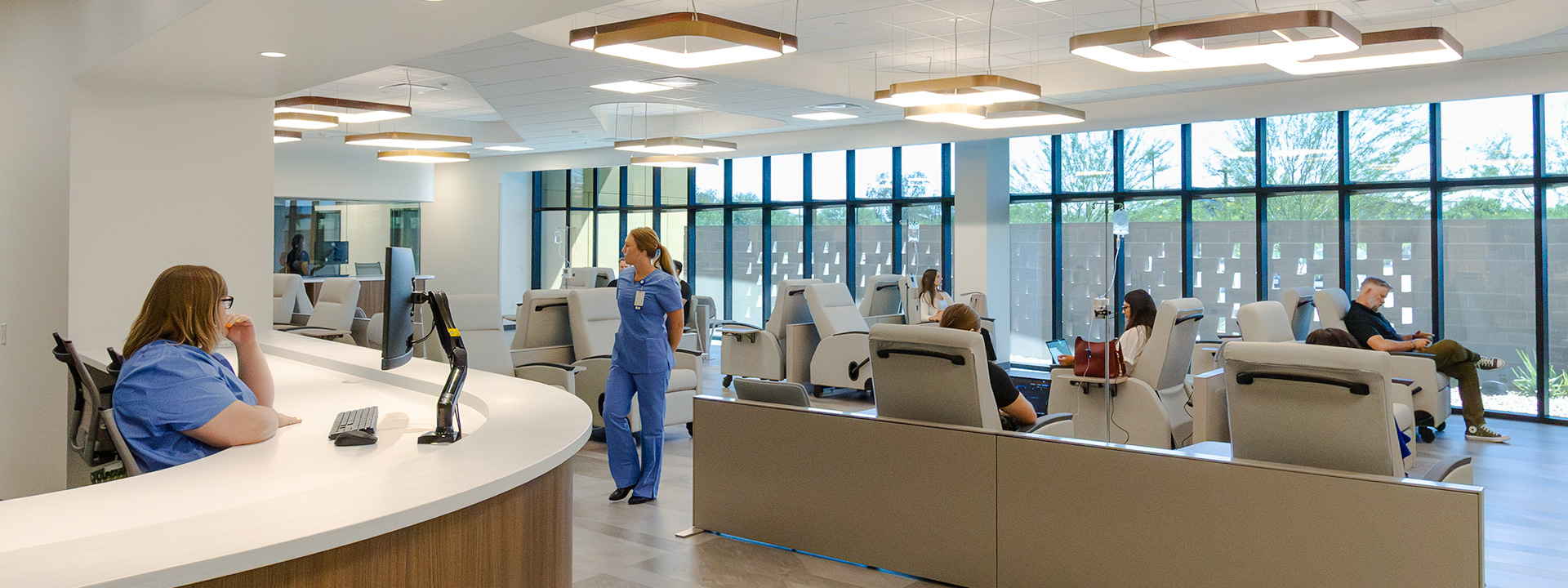 Healthcare workers and patients in a modern infusion center, with a nurse at the reception desk and patients receiving treatment in private treatment areas.